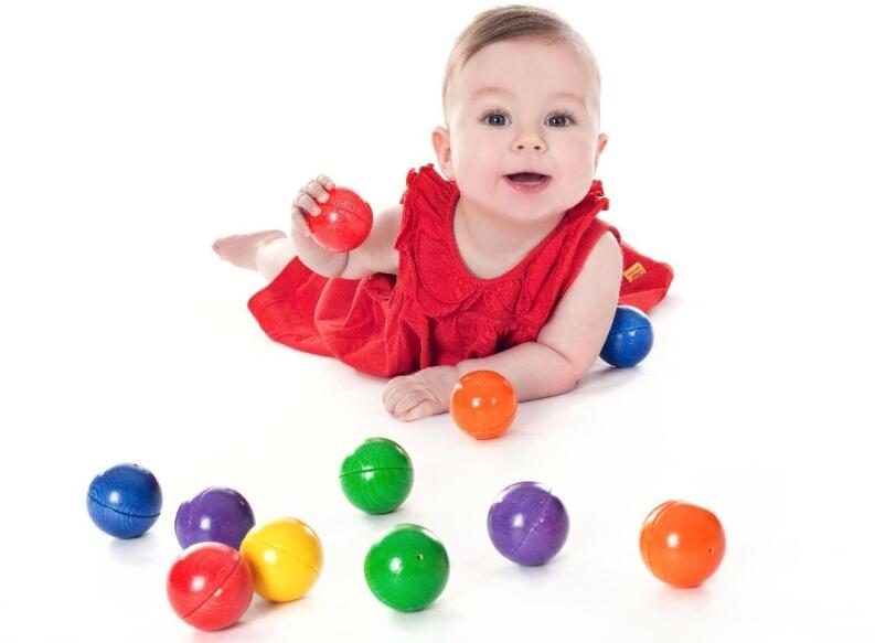 Baby in rotem Kleid spielt mit bunten Kugeln, hält eine rote Kugel in der Hand, weitere farbige
